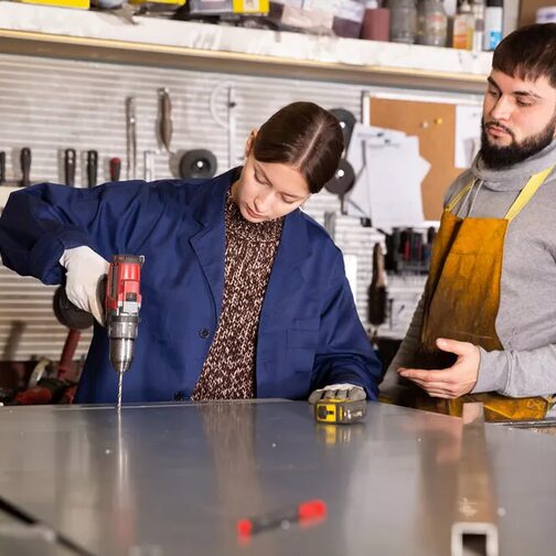 A young woman is using a power tool on a metal plate, being watched by a bearded man.