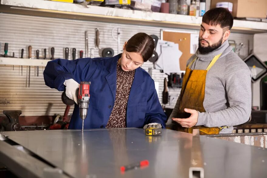 A young woman is using a power tool on a metal plate, being watched by a bearded man.